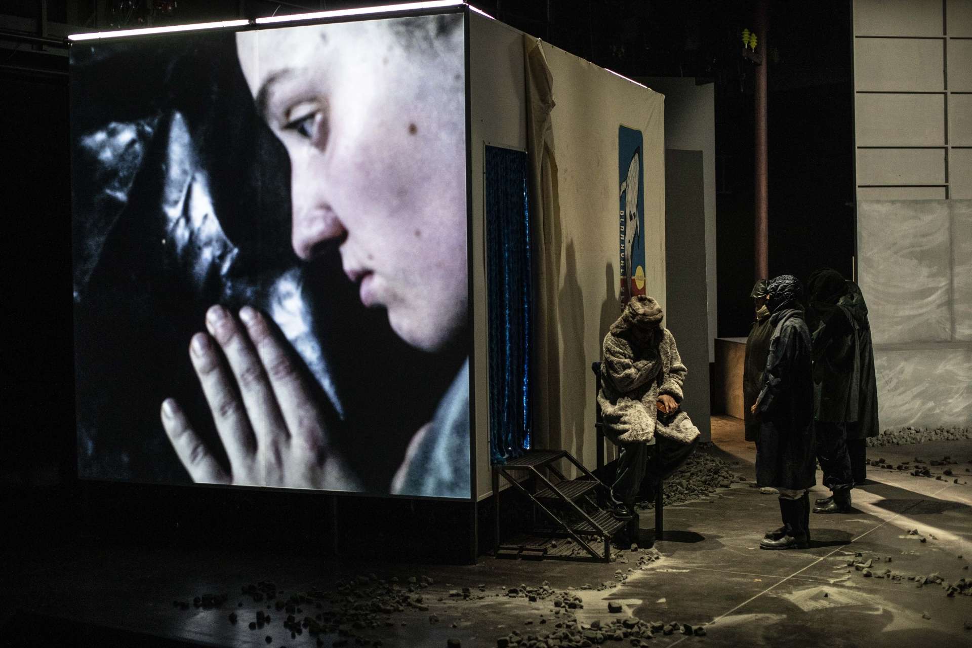 Est projetée sur un mur une vidéo où apparaît une femme, sa main et son visage couché sur une surface de cuir. À côté d'un autre mur, un groupe de personnes habillées d'habits noirs cachant leurs identités se tiennent face à une personne portants des vêtements de fourrure.
