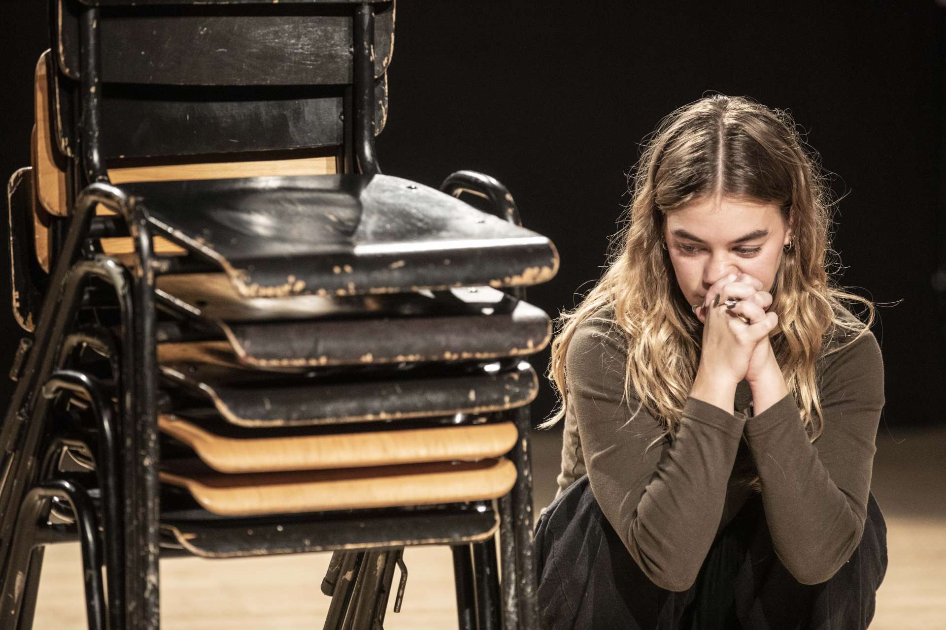 Groupe 50 élèves en atelier jeu avec Emmanuel Matte photo Jean-Louis Fernandez une personnes assise au sol à coté d'une pile de chaises en exercice d'interpretation les doigts croisés ramenés au visages pensive